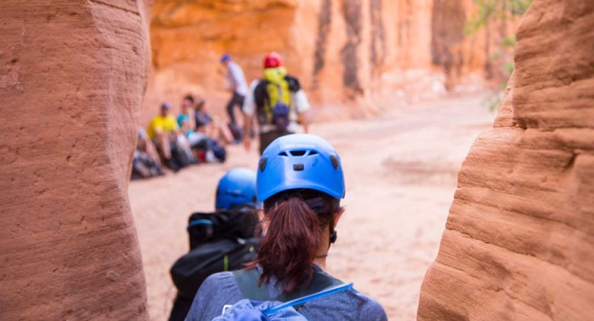 Two people wearing helmets exit a slot canyon toward a group of people.